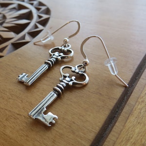 May include: A pair of silver key-shaped earrings with ornate details. The earrings are on a wooden surface.