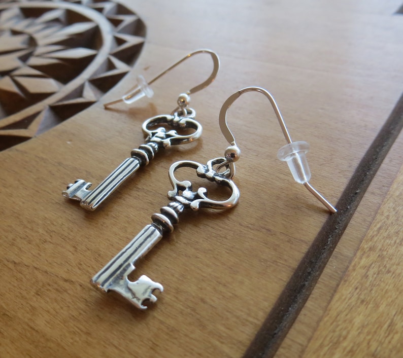 May include: A pair of silver key-shaped earrings with ornate details. The earrings are on a wooden surface.