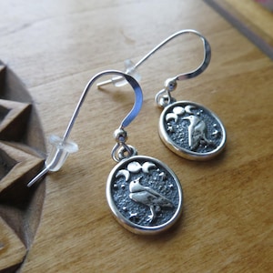 May include: Silver earrings with a crescent moon and raven design. The raven is perched on a branch with stars in the background.