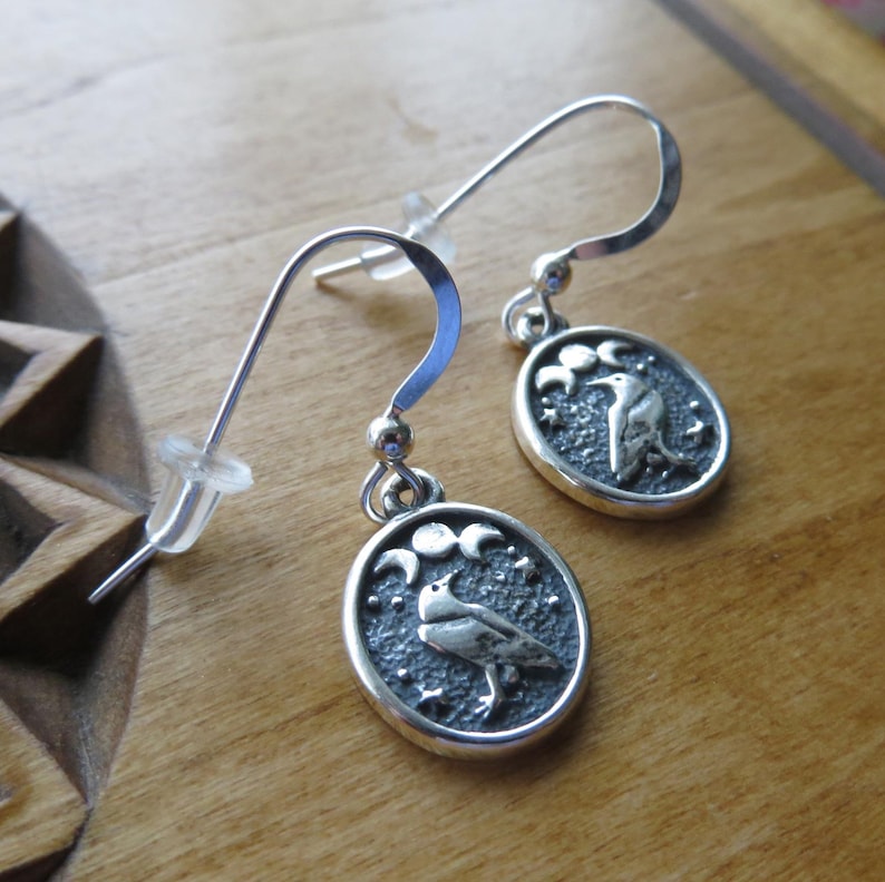 May include: Silver earrings with a crescent moon and raven design. The raven is perched on a branch with stars in the background.