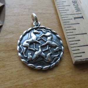 May include: Silver pendant featuring a circular design with four horses running in a circle. The horses are in a stylized design and are facing inwards towards the center of the circle. The pendant is on a wooden surface.