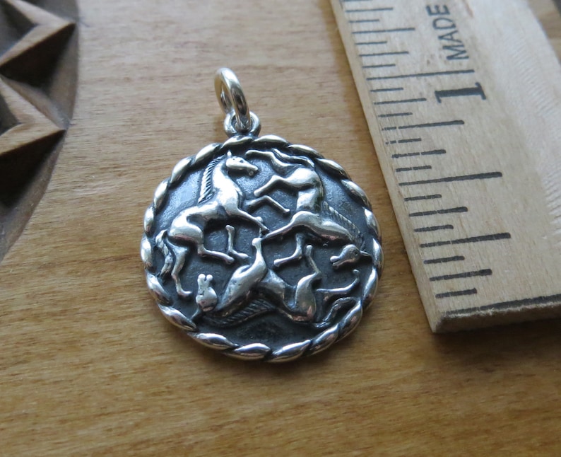 May include: Silver pendant featuring a circular design with four horses running in a circle. The horses are in a stylized design and are facing inwards towards the center of the circle. The pendant is on a wooden surface.
