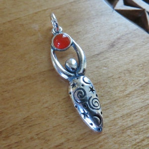 May include: Silver pendant featuring a stylized female figure holding a round carnelian gemstone. The figure is adorned with swirling designs and stars.