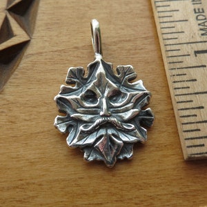 Op de afbeelding: Een zilveren hanger met een gedetailleerd groen man gezicht met een baard en snor, omringd door bladeren. De hanger is ongeveer 2,5 cm in diameter.