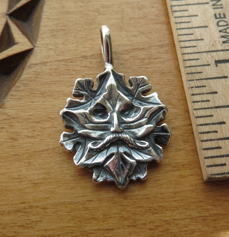 Op de afbeelding: Een zilveren hanger met een gedetailleerd groen man gezicht met een baard en snor, omringd door bladeren. De hanger is ongeveer 2,5 cm in diameter.