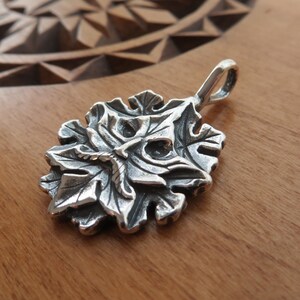 Op de afbeelding: Een zilveren hanger in de vorm van een gestileerd gezicht met bladeren, die lijkt op een groene man. De hanger heeft een lus aan de bovenkant om aan een ketting te bevestigen.