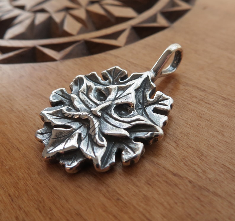 Op de afbeelding: Een zilveren hanger in de vorm van een gestileerd gezicht met bladeren, die lijkt op een groene man. De hanger heeft een lus aan de bovenkant om aan een ketting te bevestigen.