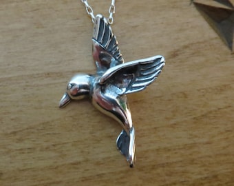 May include: A silver hummingbird pendant necklace. The hummingbird is in flight with its wings spread out. The necklace is on a wooden surface.