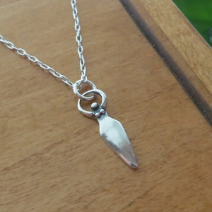 May include: A silver necklace with a pendant shaped like a stylized female figure. The pendant has a circle around the figure's head and a small bead at the bottom.