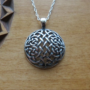 May include: A circular silver pendant featuring an intricate, openwork interlaced knot pattern. The pendant is suspended from a matching silver chain, showcasing a polished finish with darkened recessed areas that highlight the detailed metalwork.