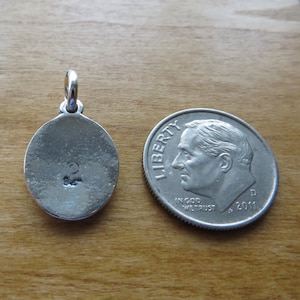 May include: A silver oval pendant with a small inscription on the back. The pendant is next to a US dime for size comparison.