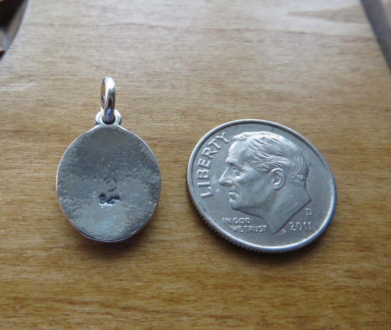 May include: A silver oval pendant with a small inscription on the back. The pendant is next to a US dime for size comparison.