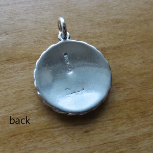 May include: The back of a silver pendant with a scalloped edge. The pendant is marked "Sterling" and has a signature.