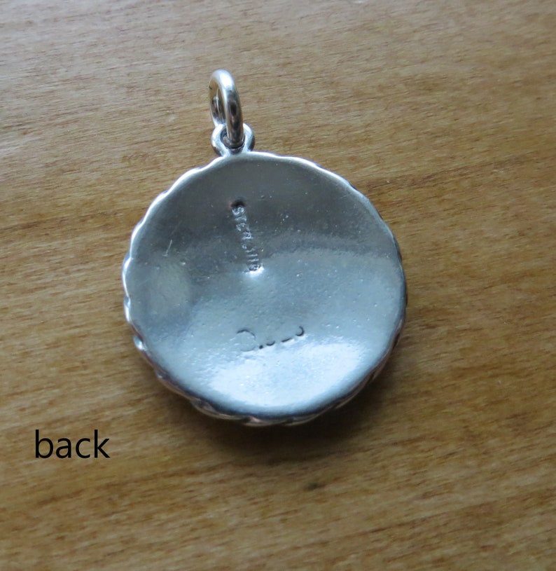May include: The back of a silver pendant with a scalloped edge. The pendant is marked "Sterling" and has a signature.