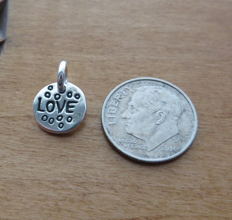 Może przedstawiać: Mała, okrągła zawieszka z metalu w kolorze srebrnym z napisem LOVE wytłoczonym czarnymi literami, otoczonym małymi wypukłymi kropkami. Zawieszka posiada k&oacute;łeczko u g&oacute;ry i znajduje się obok amerykańskiej monety dziesięciocentowej dla skali.