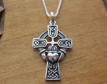 Puede incluir: Un colgante de plata en forma de cruz celta con un diseño de corazón y corona. La cruz está detallada con intrincados nudos.