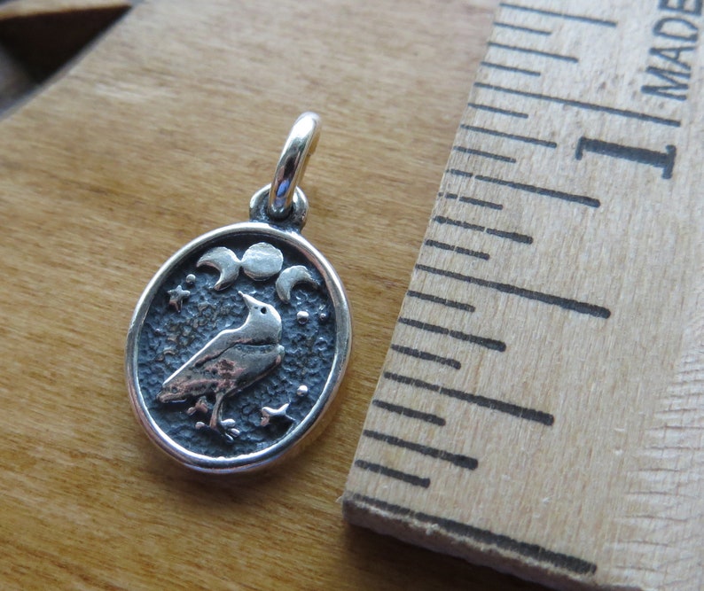 May include: Silver charm featuring a stylized raven with a crescent moon and stars. The charm is oval shaped and has a textured surface.