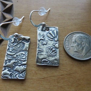 May include: A pair of silver earrings with a textured design. One earring features a seashell design, while the other has a more abstract pattern. The earrings are shown next to a US dime for scale.