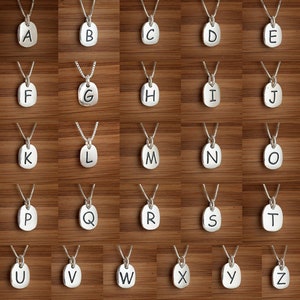 Puede incluir: Un conjunto de 26 collares de plata, cada uno con un dije con forma de letra. Los dijes son de forma ovalada y tienen una superficie ligeramente texturizada. Las letras est&aacute;n impresas en negro.
