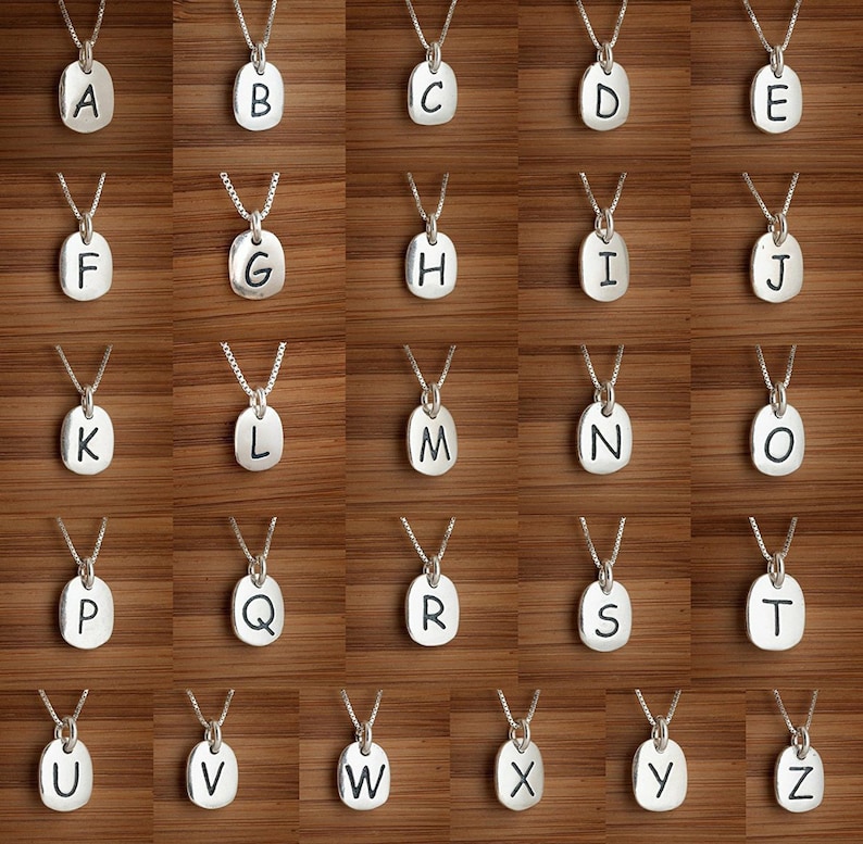 Puede incluir: Un conjunto de 26 collares de plata, cada uno con un dije con forma de letra. Los dijes son de forma ovalada y tienen una superficie ligeramente texturizada. Las letras est&aacute;n impresas en negro.