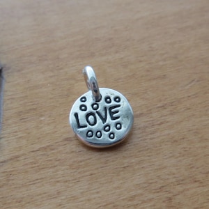 Może przedstawiać: Mała, okrągła zawieszka z metalu w kolorze srebrnym z wytłoczonym na środku słowem LOVE czarnymi wielkimi literami, otoczona serią małych, wypukłych kropek, z k&oacute;łeczkiem przymocowanym u g&oacute;ry w celu zawieszenia.
