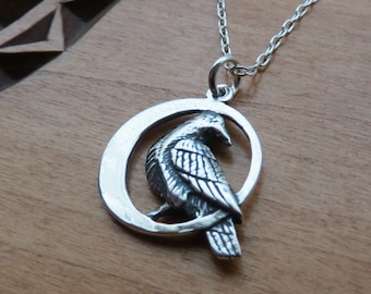 May include: A silver necklace with a pendant featuring a detailed raven perched inside a crescent moon shape.