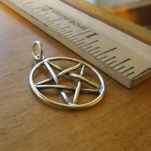 May include: A silver pendant in the shape of a pentagram, or five-pointed star, with a circular border. The pendant is on a wooden surface.