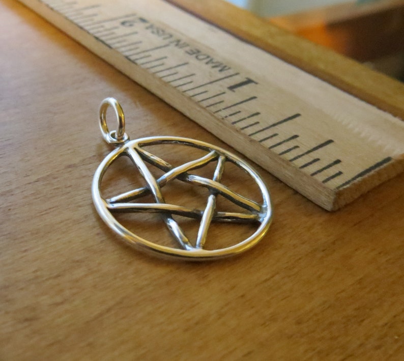 May include: A silver pendant in the shape of a pentagram, or five-pointed star, with a circular border. The pendant is on a wooden surface.