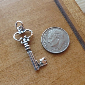 May include: A silver key charm with an ornate design. The key is about the size of a dime.
