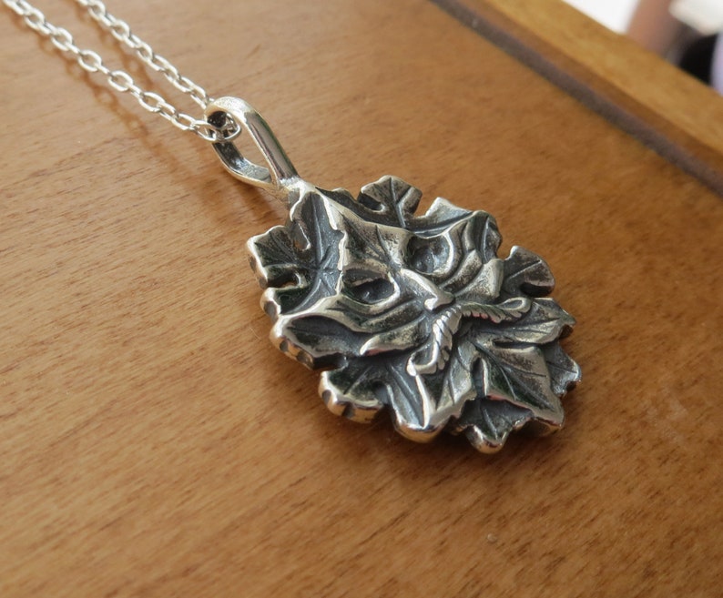 Op de afbeelding: Een zilveren ketting met een hanger met een gedetailleerd, driedimensionaal ontwerp van een gezicht omringd door bladeren.