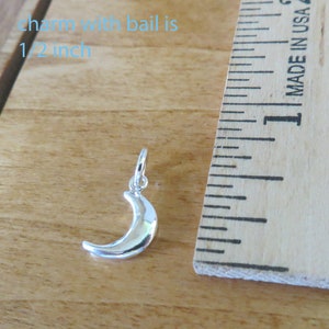 May include: A silver crescent moon charm with a bail, measuring 1/2 inch in size. The charm is shown next to a ruler for size reference.