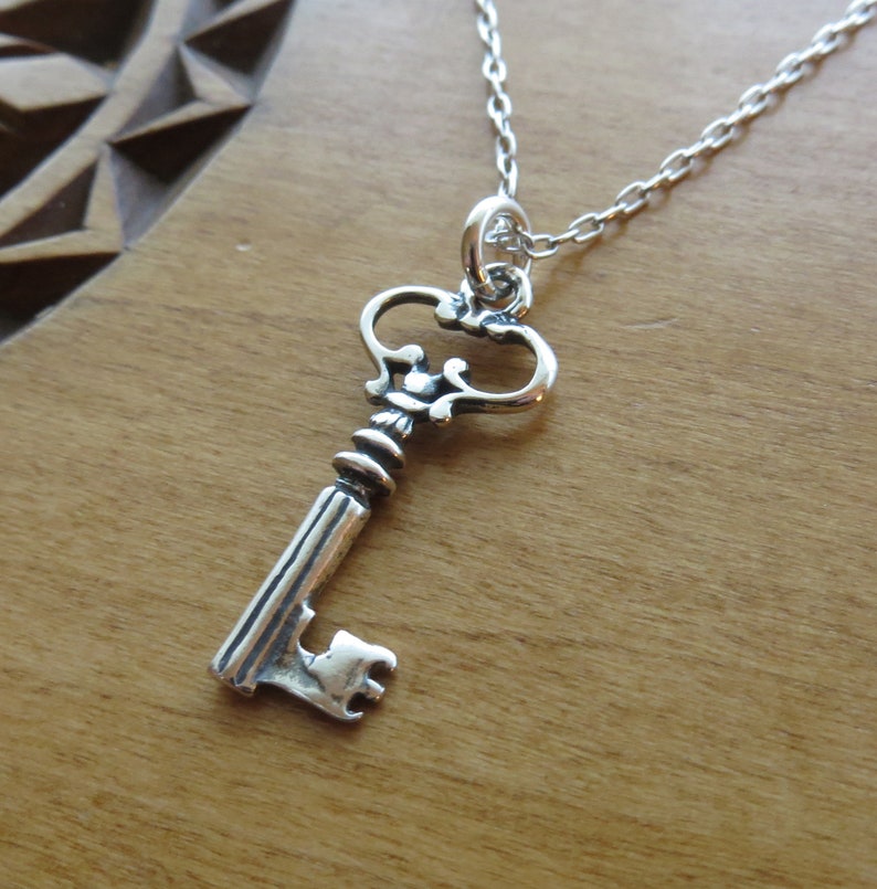 May include: A silver key pendant necklace with a detailed ornate design. The key has a heart-shaped bow and a long, slender shaft.