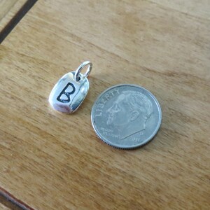 Puede incluir: Encanto de plata con la letra "B" estampada en &eacute;l. El encanto es aproximadamente del tama&ntilde;o de una moneda de diez centavos.