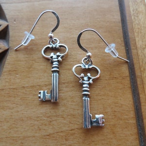 May include: A pair of silver key-shaped earrings with intricate details. The keys have a vintage style and are suspended from silver hooks.