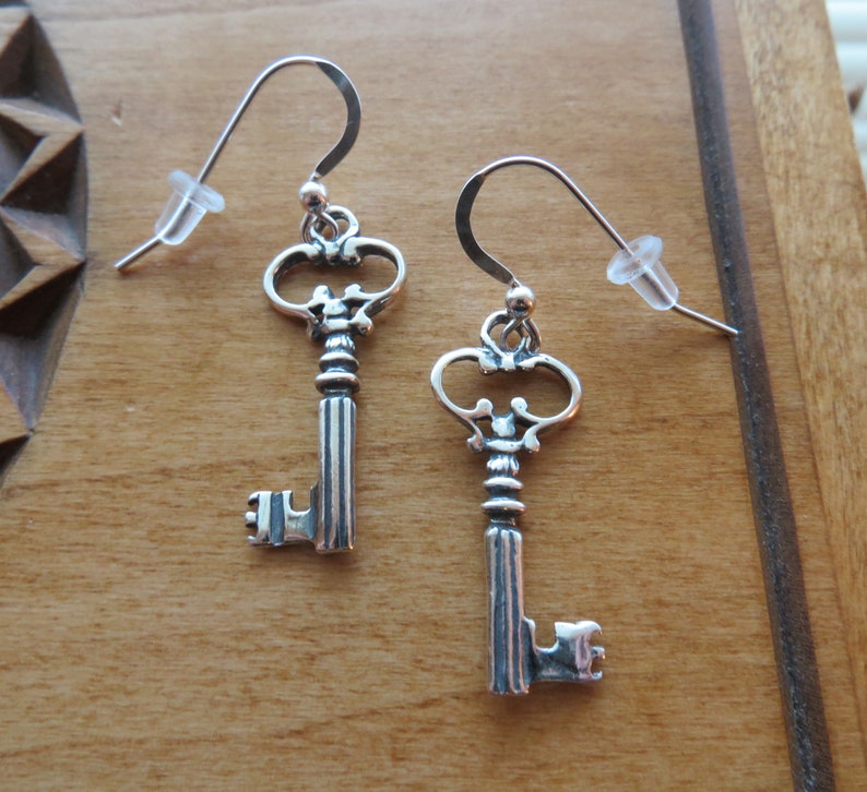 May include: A pair of silver key-shaped earrings with intricate details. The keys have a vintage style and are suspended from silver hooks.
