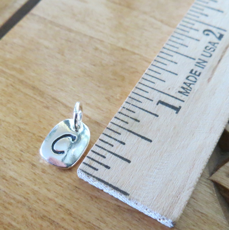 Puede incluir: Un charm de plata con la letra "C" grabada en &eacute;l. El charm mide aproximadamente 2,5 cm de largo y se muestra junto a una regla.