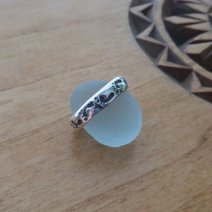 May include: A silver ring with a heart design. The ring is resting on a smooth, light blue stone. The background is a wooden surface with a carved design.
