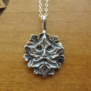Op de afbeelding: Een zilveren ketting met een gedetailleerd groen mannetje-hanger. De hanger is gevormd als een blad met een gezicht in het midden. Het gezicht heeft een baard en snor, en de ogen zijn groot en rond.