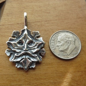 Op de afbeelding: Een zilveren hanger met een gedetailleerd groen man gezicht omringd door bladeren. De hanger is ongeveer zo groot als een Nederlandse tien-cent munt.