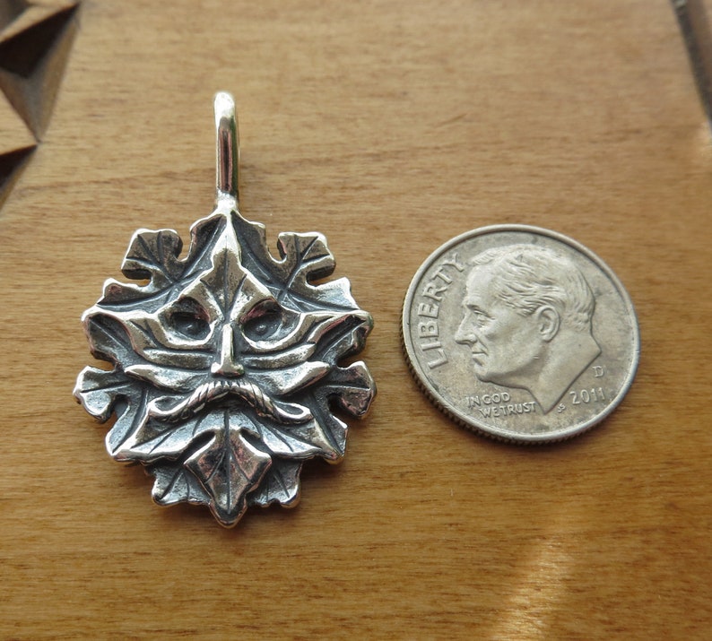 Op de afbeelding: Een zilveren hanger met een gedetailleerd groen man gezicht omringd door bladeren. De hanger is ongeveer zo groot als een Nederlandse tien-cent munt.
