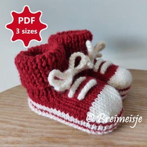 以下が含まれることがあります： 赤と白のニット製のベビーブーティで、スニーカーの形をしています。白い靴ひもが付いています。ブーティには「PDF 3サイズ」のラベルが付いています。