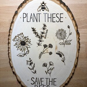 May include: A wooden slice with the text "Plant These" and "Save the Bees" burned into the surface. The slice features illustrations of various flowers, including sunflower, sage, verbena, poppy, lavender, aster, and rosemary.
