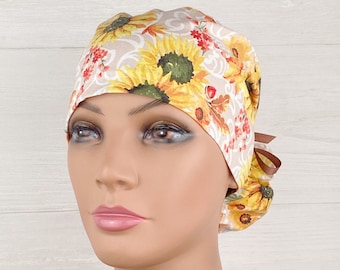 May include: A white scrub hat with a sunflower and leaf pattern. The hat has a brown tie at the back.