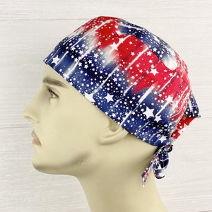 May include: A scrub cap featuring a patriotic pattern with red, white, and blue colors. The fabric displays a design of white stars scattered over a background of vertical red and blue stripes. The cap includes ties at the back for adjustment.