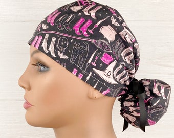 Peut inclure: Une charlotte de bloc opératoire noire ornée d'un motif répétitif d'illustrations de sorcellerie en rose et blanc, incluant des chats, chapeaux, bottes, chaudrons et boules de cristal. L'arrière est froncé et maintenu par un ruban noir.