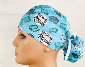 May include: A light blue surgical scrub hat with a white tie. The hat has a repeating pattern of turquoise rolled towels and the text "They See Me Rolling".