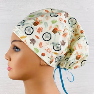 May include: A white scrub cap with a fall-themed print of bicycles, pumpkins, and leaves. The cap has a blue ribbon tie.