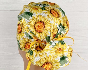 May include: A white scrub hat with a sunflower print and yellow ties. The hat is tied in a knot at the back of the head.