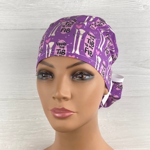 May include: Purple surgical scrub hat with a white elastic band. The hat has a repeating pattern of white bones with pink hearts and the text "You're the Tib to my Fib".