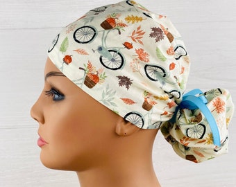 May include: A scrub cap featuring a cream-colored background with a repeating pattern of bicycles, autumn leaves, and pumpkins. The cap includes a gathered back section secured with a bright light blue ribbon, providing a snug fit for the wearer.